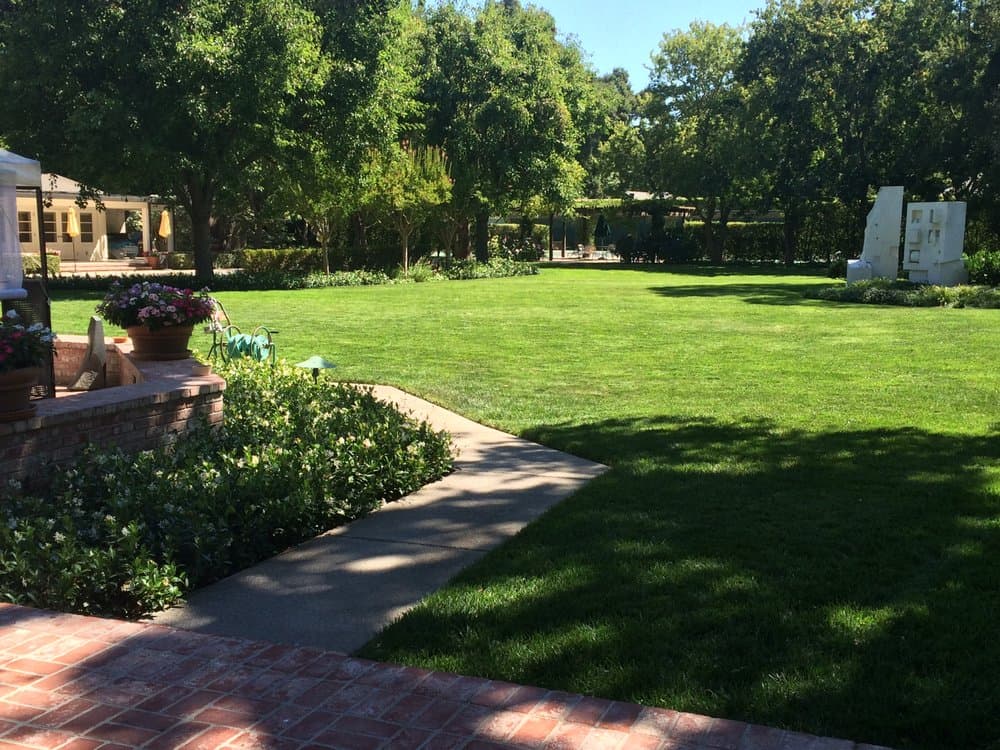 Expansive manicured estate lawn framed by mature trees, layered garden beds, and a concrete garden walkway