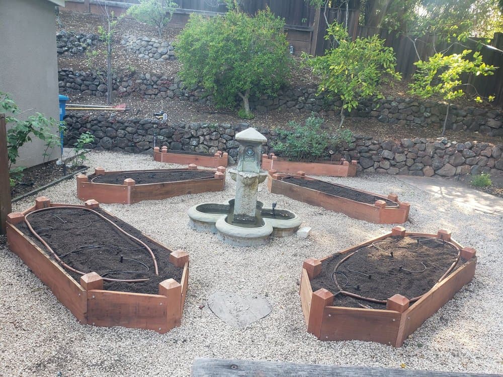 Redwood raised garden beds arranged around a central stone fountain above a stacked-stone retaining wall