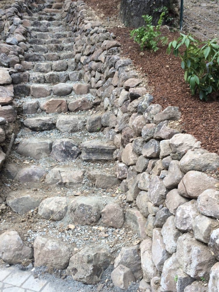Hand-set natural boulder steps climbing a slope beside a dry-stacked stone retaining wall and mulched bed